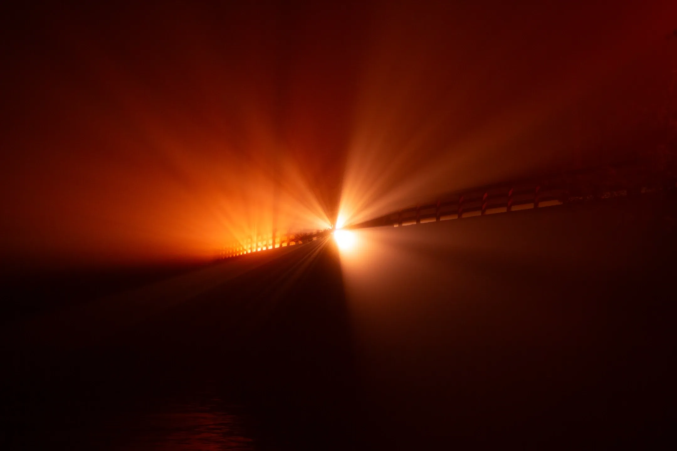 Title: Burst, Description: Bright red orange light rays shining though a fench in the fog