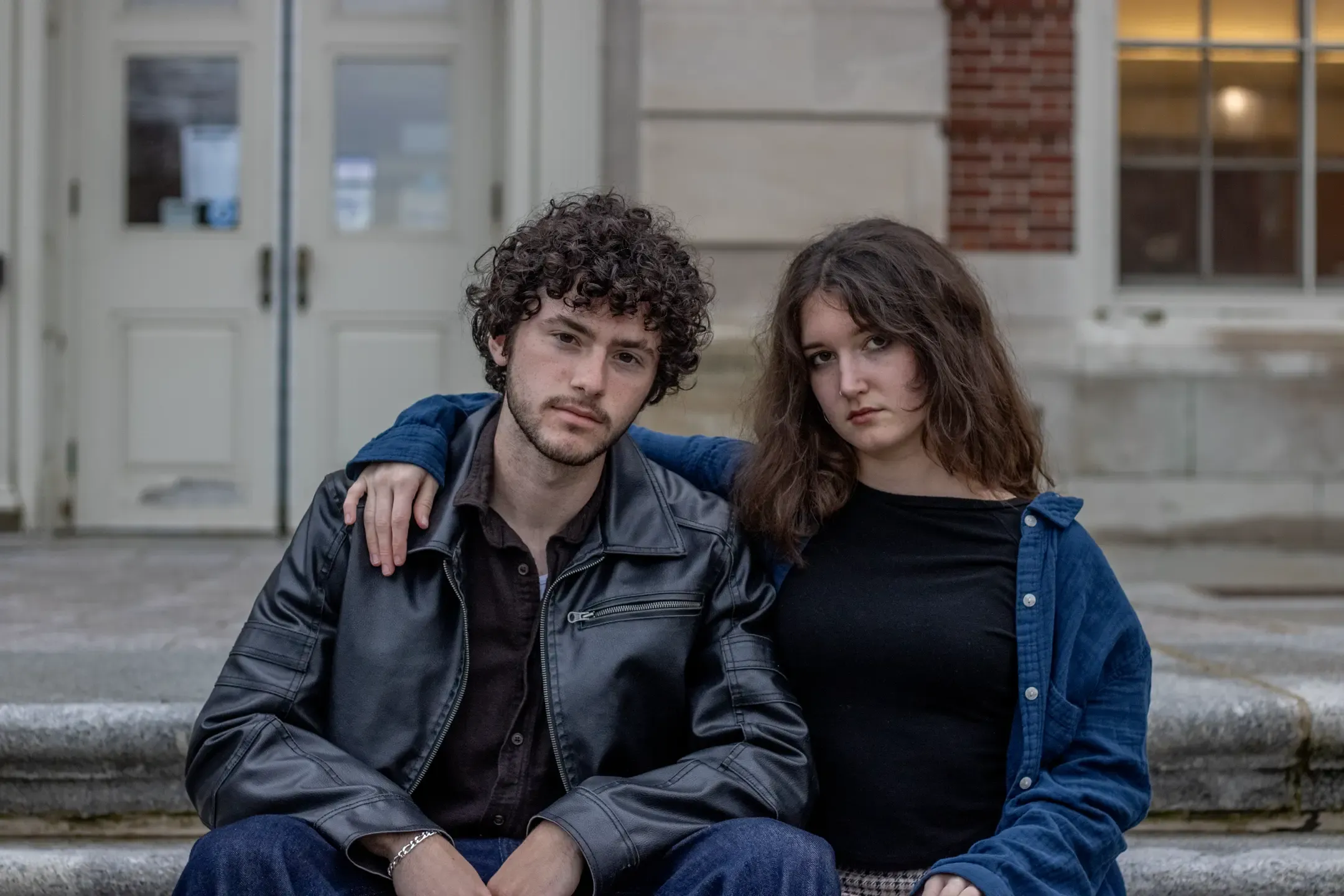 Title: Heathers, Description: A young woman with her arm over the shoulder of a young man sitting on the steps of a building